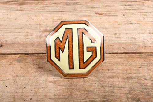 MG: An 'MG' octagonal badge, brown text and surround, cream background, mounting screw holes to rear; badge measures approximately 5 cm in diameter.