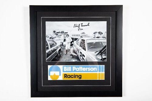 PHIL BROCK: A signed b/w photograph / display piece featuring Phil Brock c1976/77 walking around team cars, signed by Brock, 45cm high, 45cm wide overall